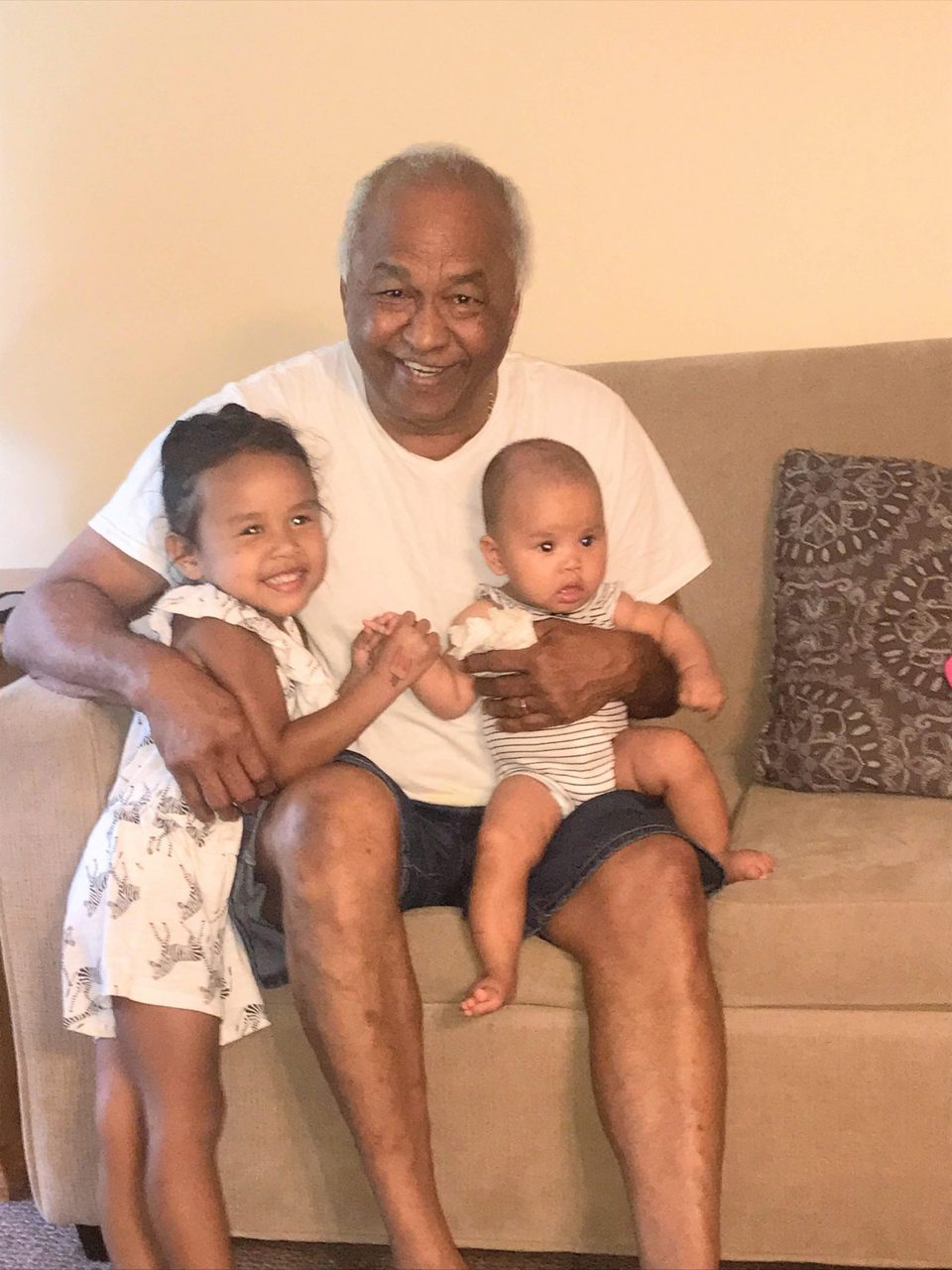 Gabriel smiling on a sofa, holding his granddaughters Kensie and baby Charli